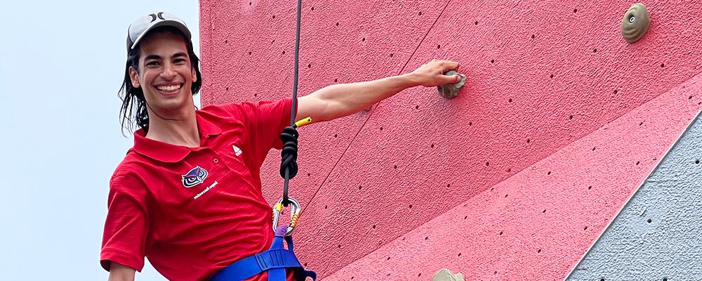 A student smiling as he hangs from the climbing wall